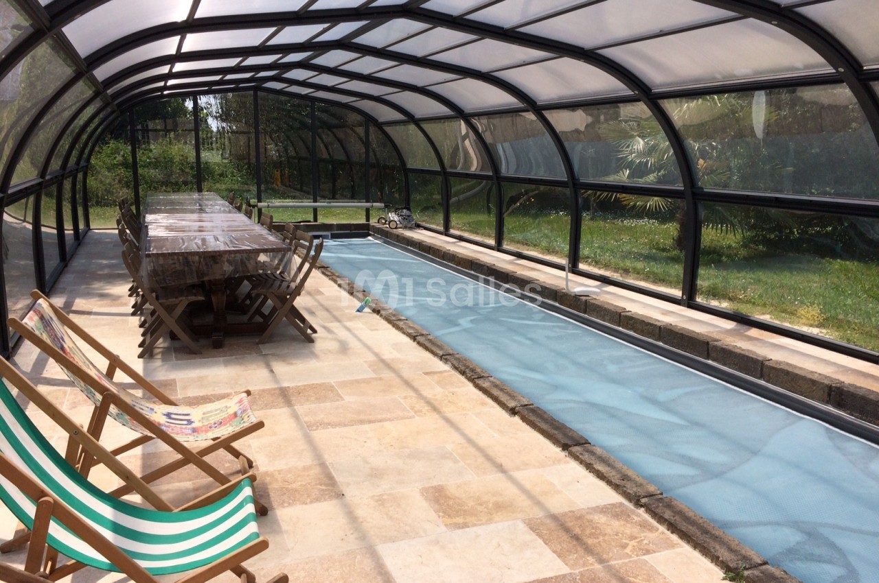 Abri transparent couvrant une piscine étroite, avec une table en bois et des transats alignés sur le côté.