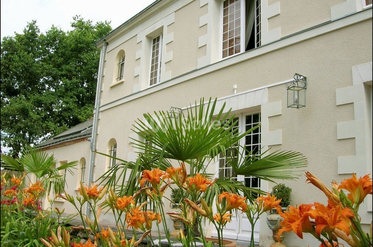 Façade d'une maison beige avec des fenêtres blanches, entourée de fleurs orange et de palmiers sous un ciel clair.
