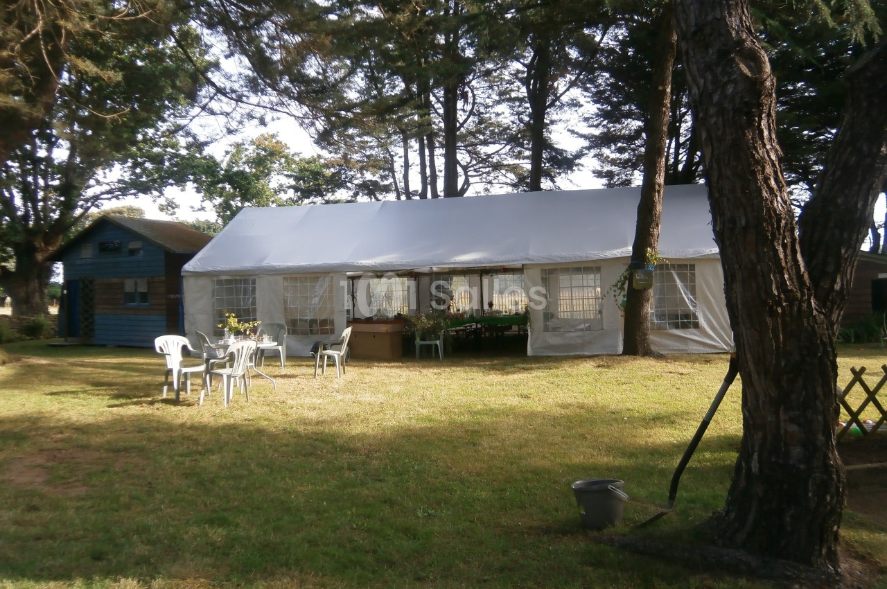 Tente blanche installée dans un jardin arboré avec des chaises en plastique et une petite cabane en bois à proximité.