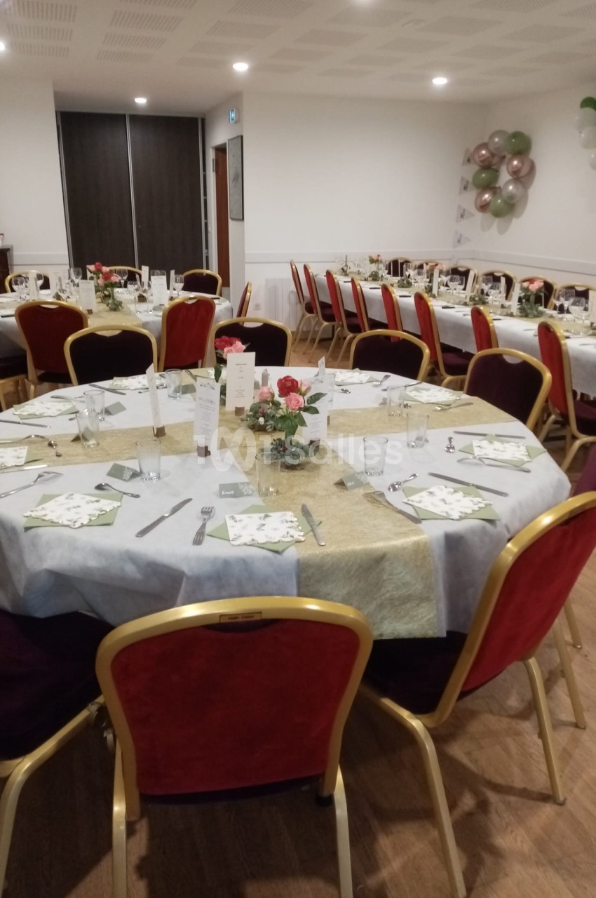 Salle de réception avec tables rondes dressées, nappes blanches et dorées, chaises rouges, décorations florales et ballons.