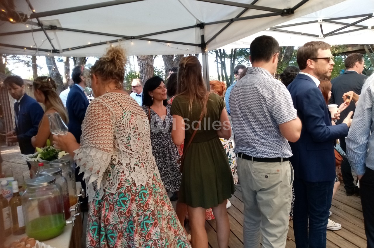 Personnes rassemblées sous une tente blanche lors d'un événement en extérieur, discutant et se servant des boissons.