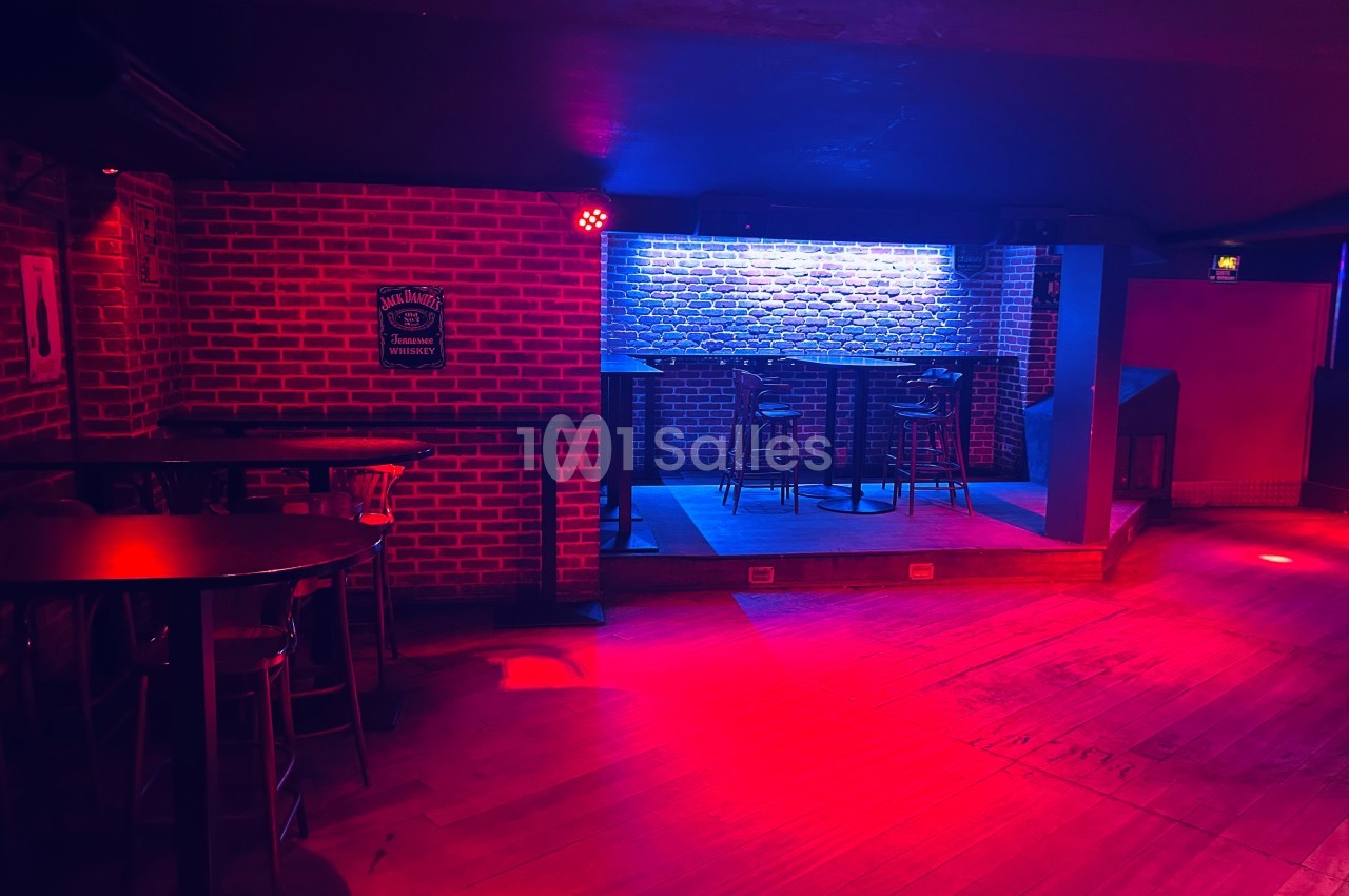 Salle de bar avec murs en briques, éclairage rouge et bleu, tables hautes et une petite scène surélevée.