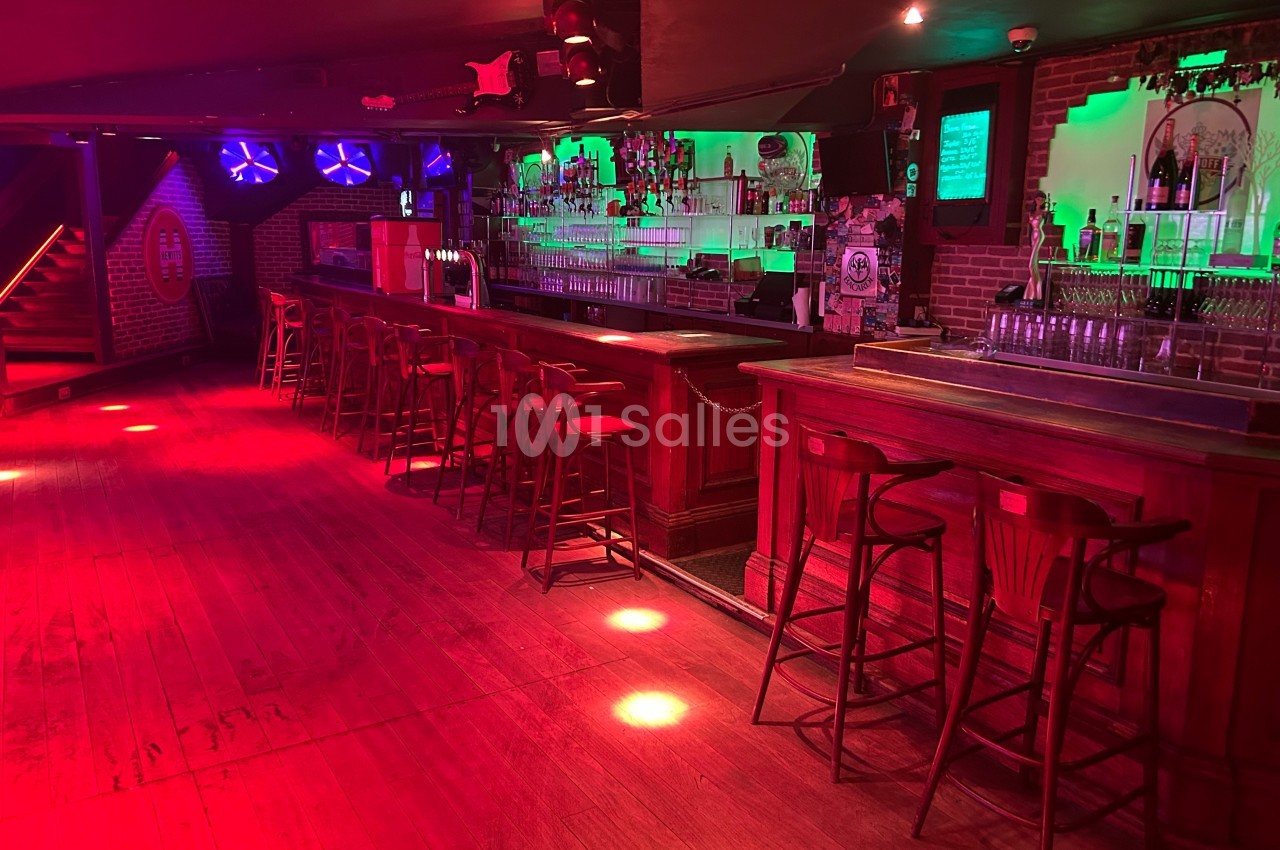 Bar avec comptoirs en bois, tabourets alignés, éclairage rouge et vert, et ambiance tamisée dans une salle vide.