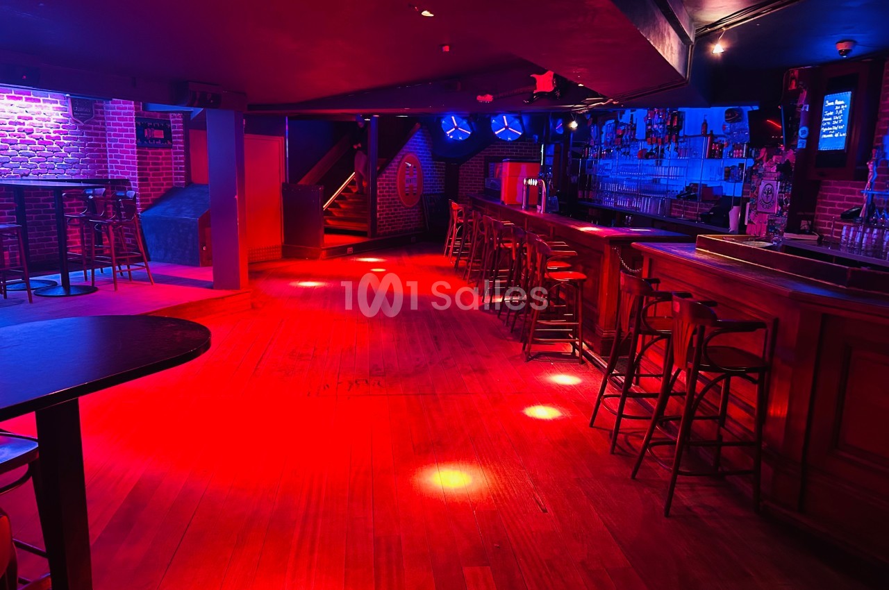 Salle de bar avec éclairage rouge tamisé, comptoir en bois, tabourets alignés et tables hautes dispersées.