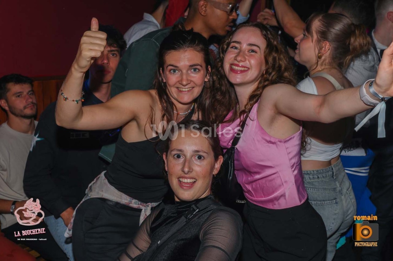 Trois jeunes femmes souriantes posent ensemble dans une ambiance festive, entourées d'autres personnes.