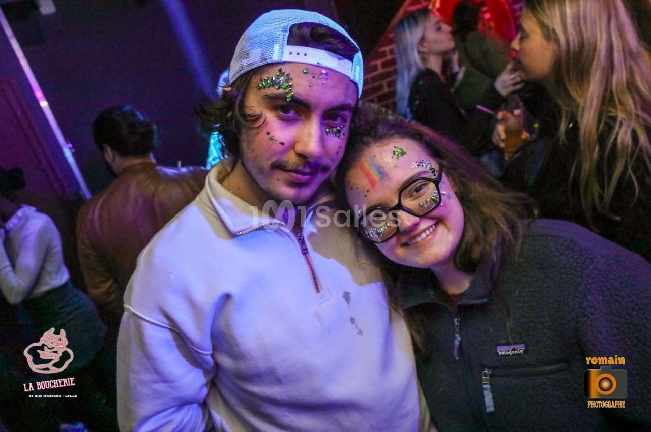 Deux personnes souriantes avec des maquillages colorés et pailletés dans un environnement festif.
