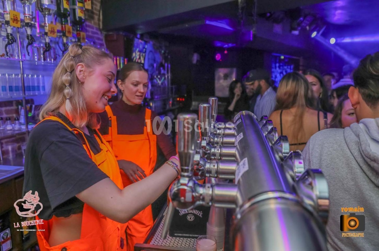 Deux serveuses en tabliers orange servent des boissons derrière un bar dans une ambiance de soirée animée.