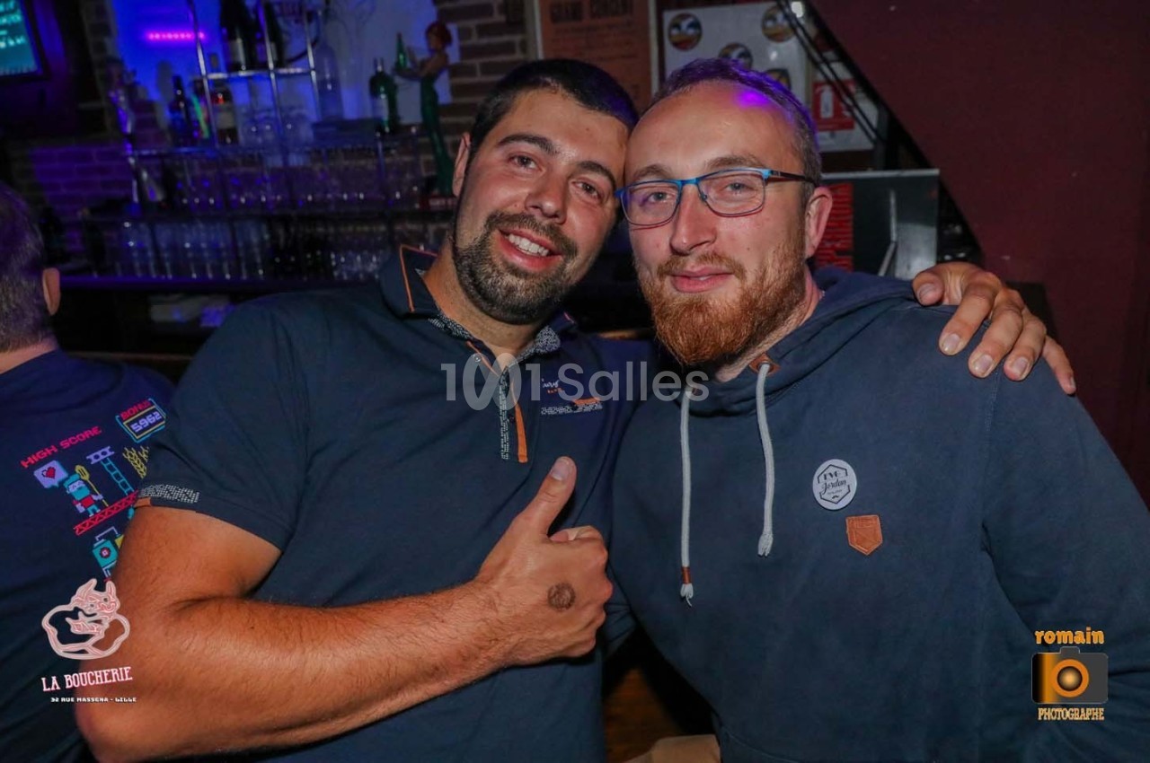 Deux hommes souriants posent côte à côte dans un bar, avec des verres et des bouteilles en arrière-plan.