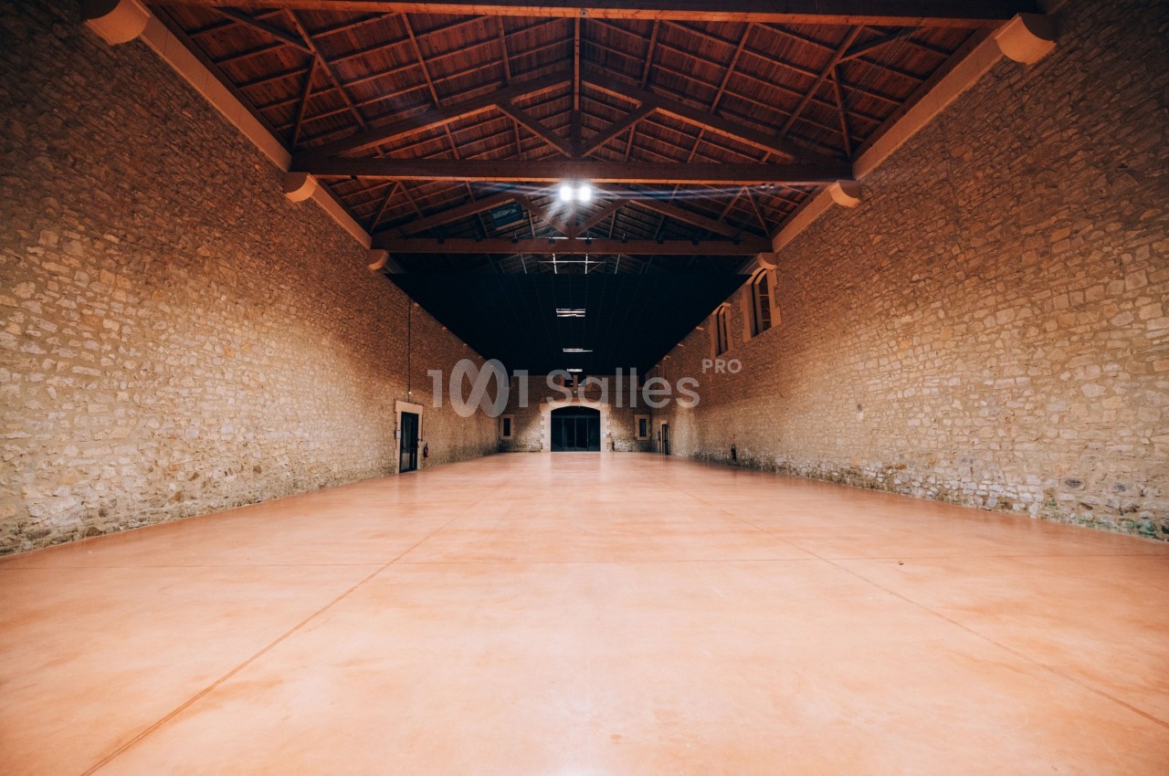 Grande salle vide avec murs en pierre, sol en béton lisse et plafond en bois avec poutres apparentes.