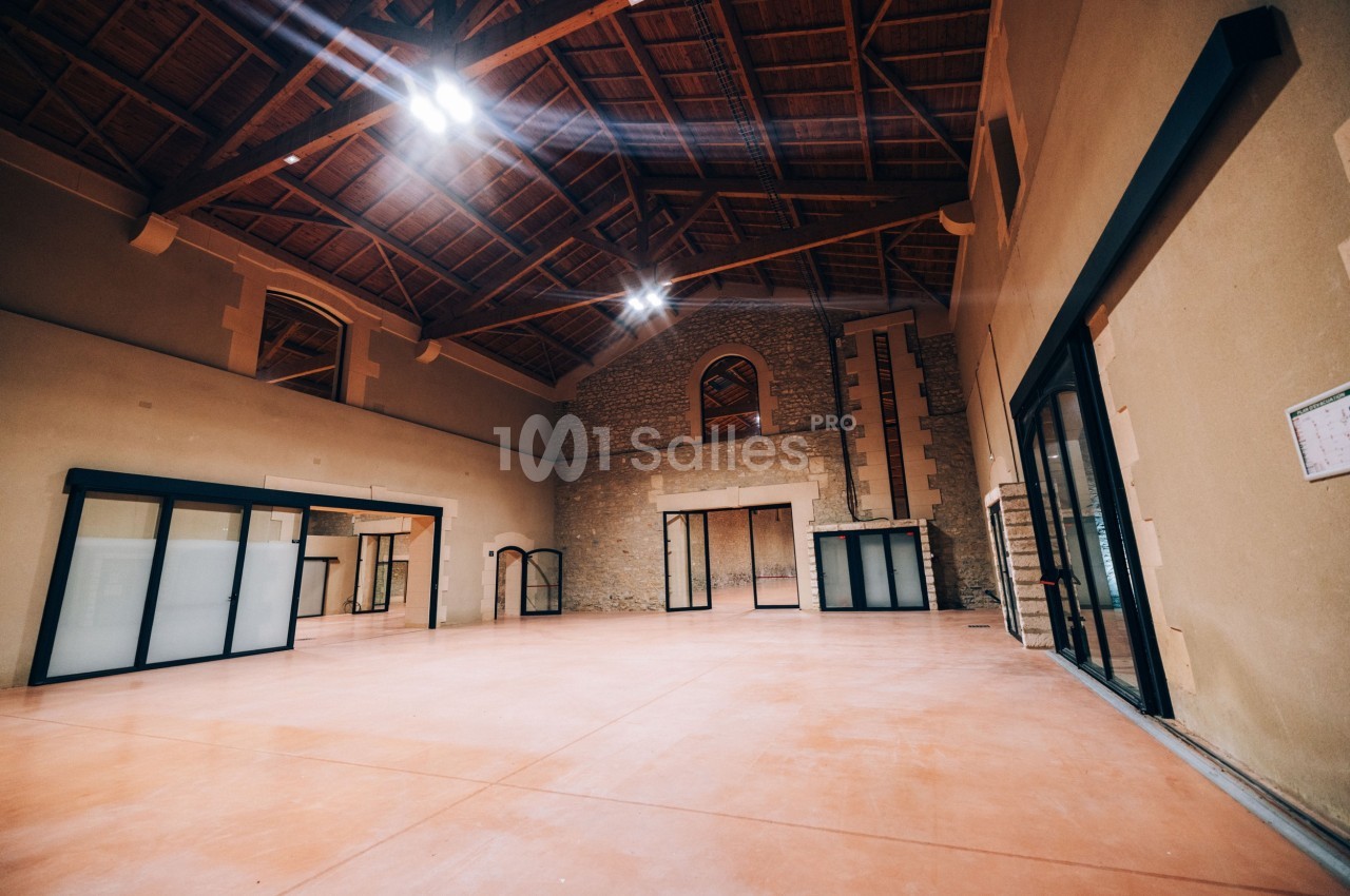 Salle spacieuse avec plafond en bois, murs en pierre et grandes baies vitrées laissant entrer la lumière naturelle.