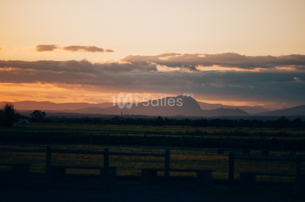 Paysage au coucher du soleil avec des montagnes à l'horizon, des champs et une clôture au premier plan.