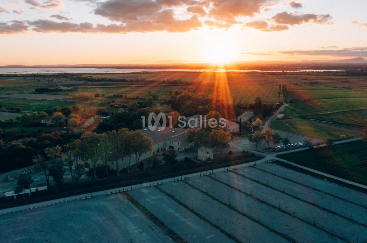 Vue aérienne d'une campagne au coucher du soleil, avec des bâtiments entourés de champs et des rayons lumineux.