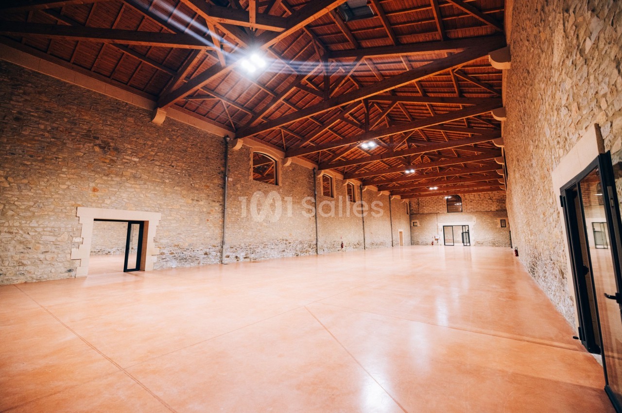 Grande salle avec murs en pierre, plafond en bois avec poutres apparentes et sol en béton poli, éclairée par des luminaires…