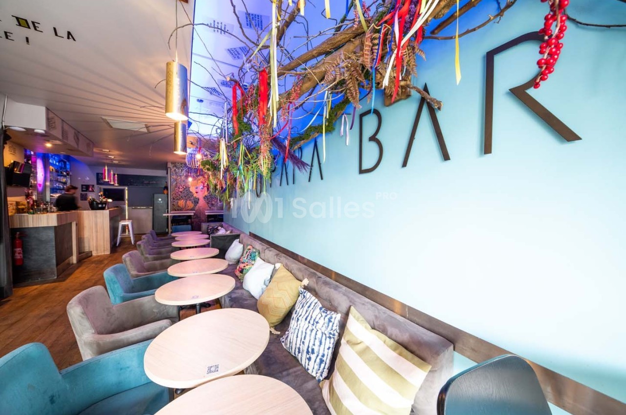 Intérieur d'un bar moderne avec tables rondes, canapés colorés et décorations suspendues sur un mur bleu.