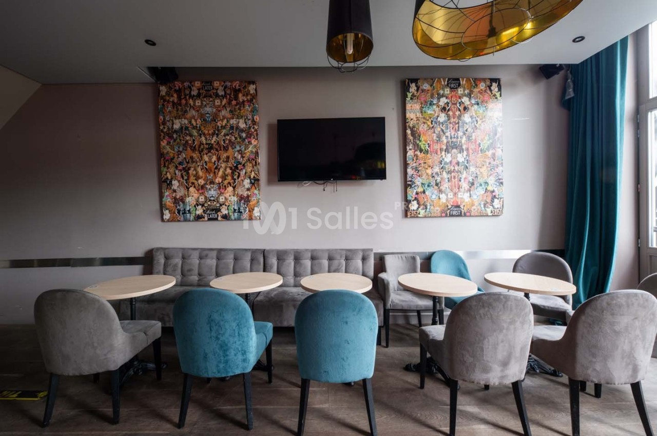 Salle moderne avec tables rondes, chaises colorées, canapé gris, télé murale et tableaux décoratifs.