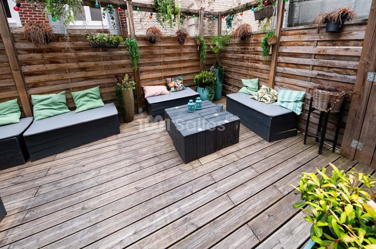 Terrasse en bois avec banquettes, coussins colorés, table basse noire et plantes suspendues et en pots.