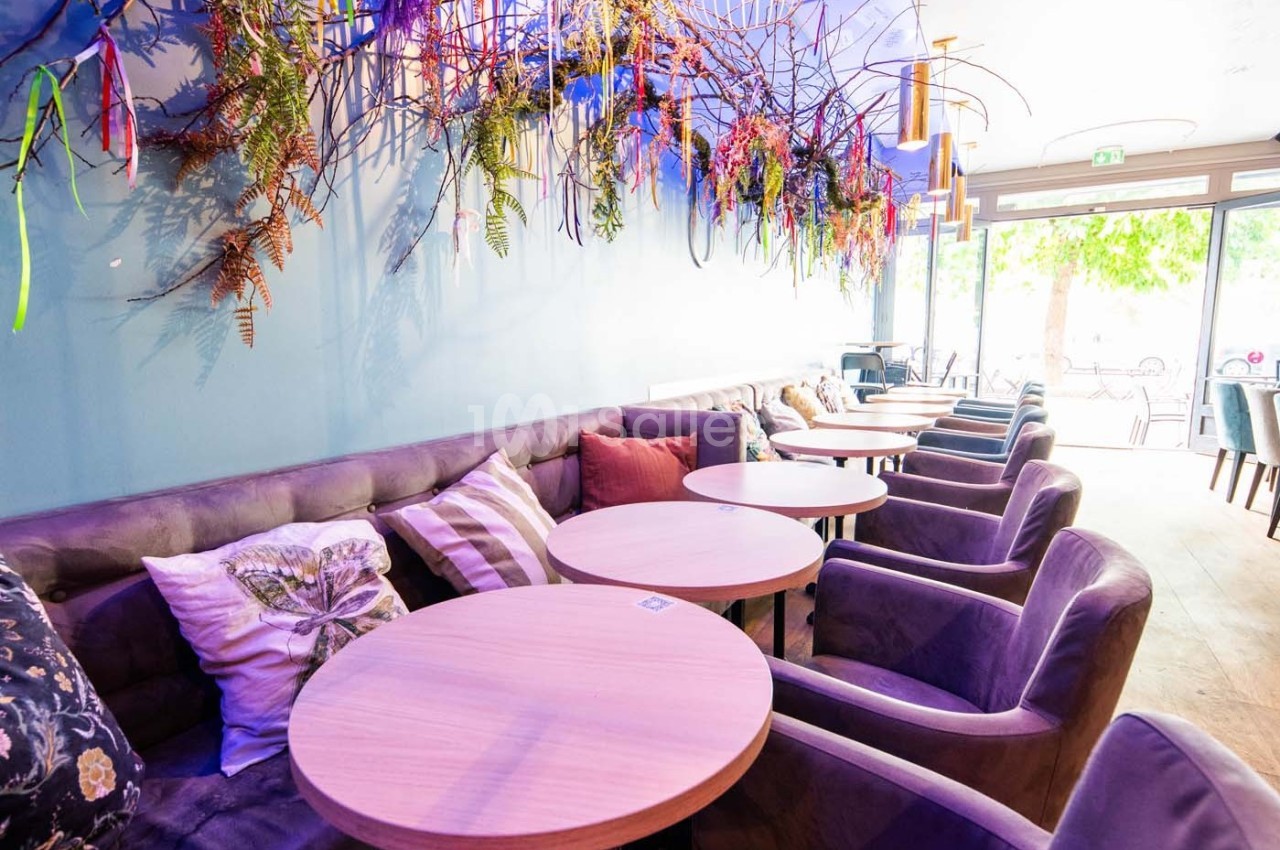 Salle lumineuse avec tables rondes en bois, fauteuils rembourrés et décoration murale végétale colorée.