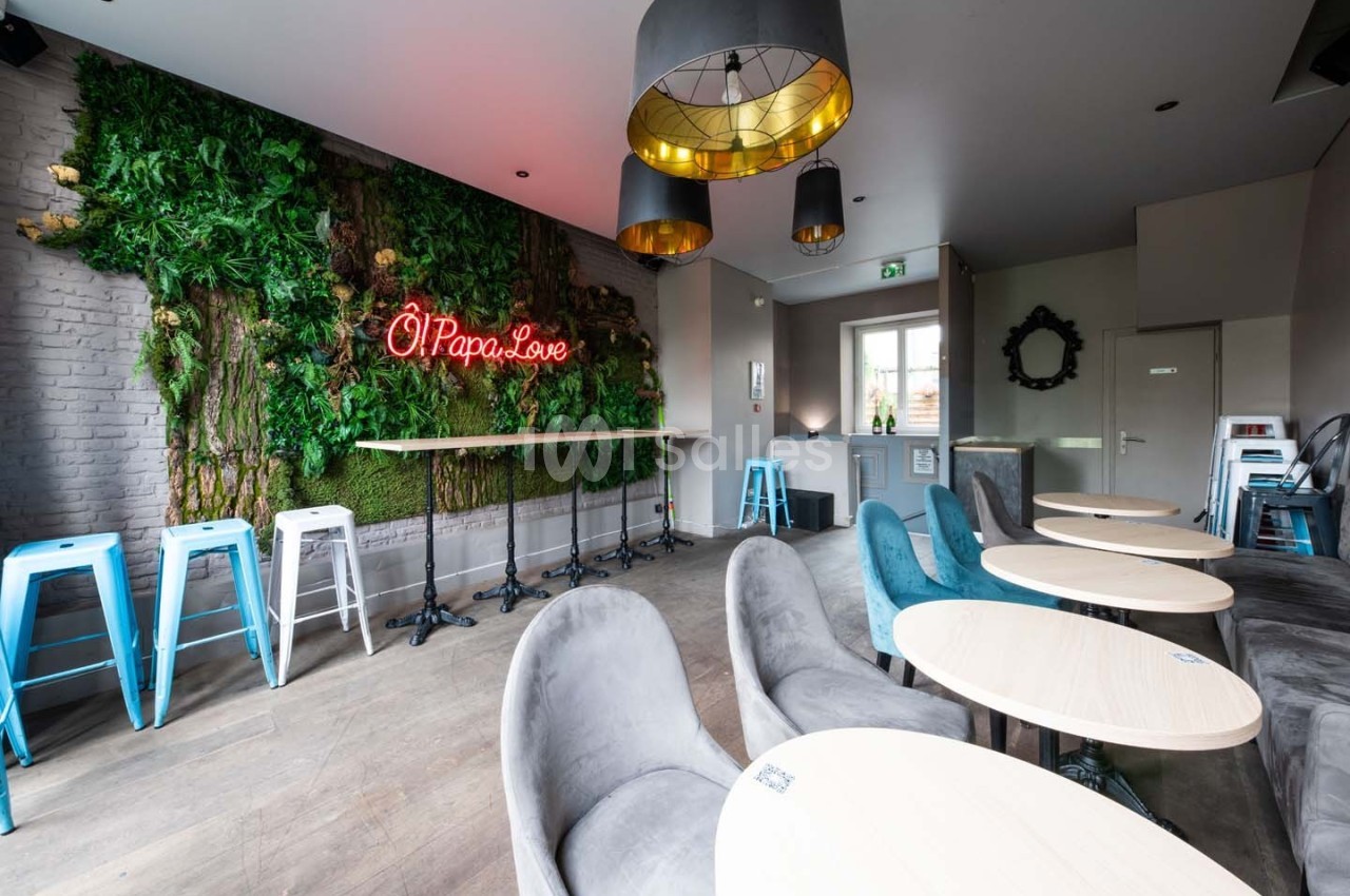 Salle moderne avec tables rondes, chaises grises, mur végétal, néon rouge ’Ô Papa Love’ et tabourets bleus près du bar.
