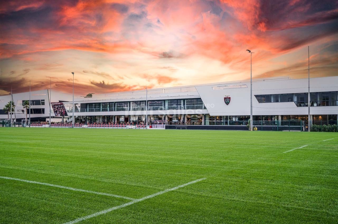 Terrain de rugby avec pelouse verte devant un bâtiment moderne sous un ciel coloré au coucher du soleil.