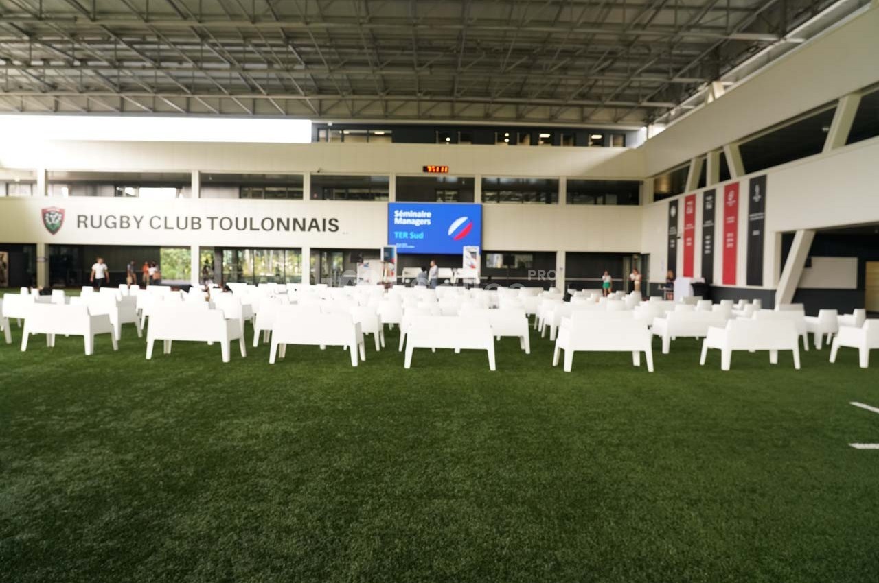 Chaises blanches disposées sur une pelouse synthétique devant un écran et des panneaux au Rugby Club Toulonnais.