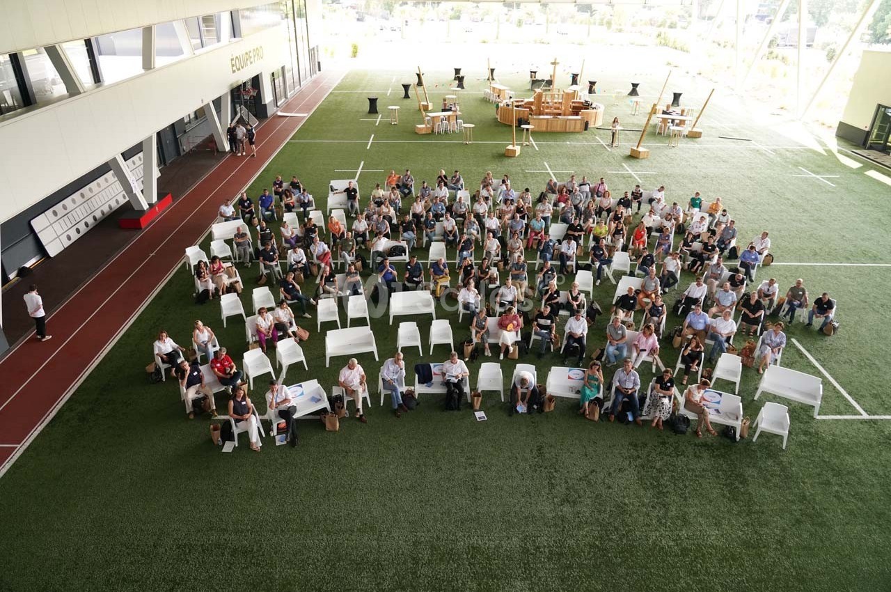 Vue aérienne d'un groupe de personnes assises sur des chaises blanches dans un espace intérieur avec sol en gazon…
