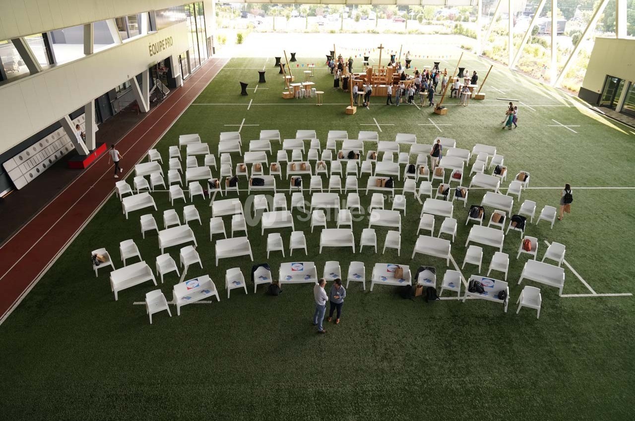Chaises blanches disposées en rangées sur un terrain synthétique, avec des tables et des personnes en arrière-plan.
