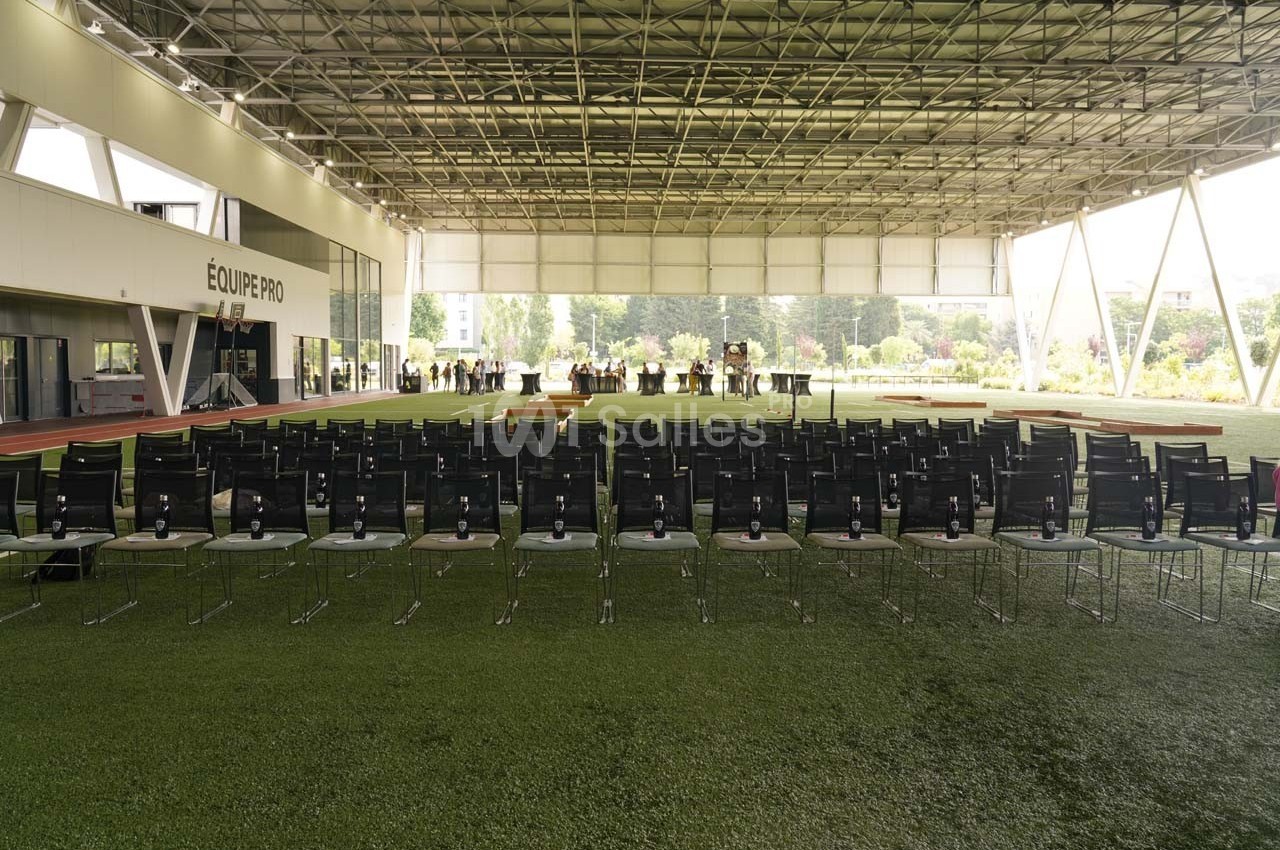 Salle de conférence en plein air avec rangées de chaises noires alignées sur un sol en gazon synthétique.