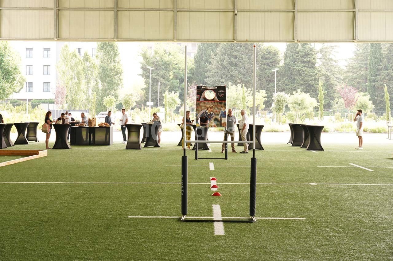 Un terrain intérieur avec des obstacles de sport, des tables hautes et des personnes en arrière-plan.