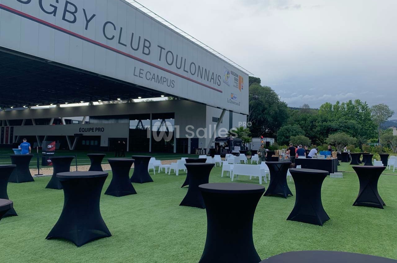 Espace extérieur avec tables hautes et chaises disposées sur une pelouse devant un bâtiment du Rugby Club Toulonnais.
