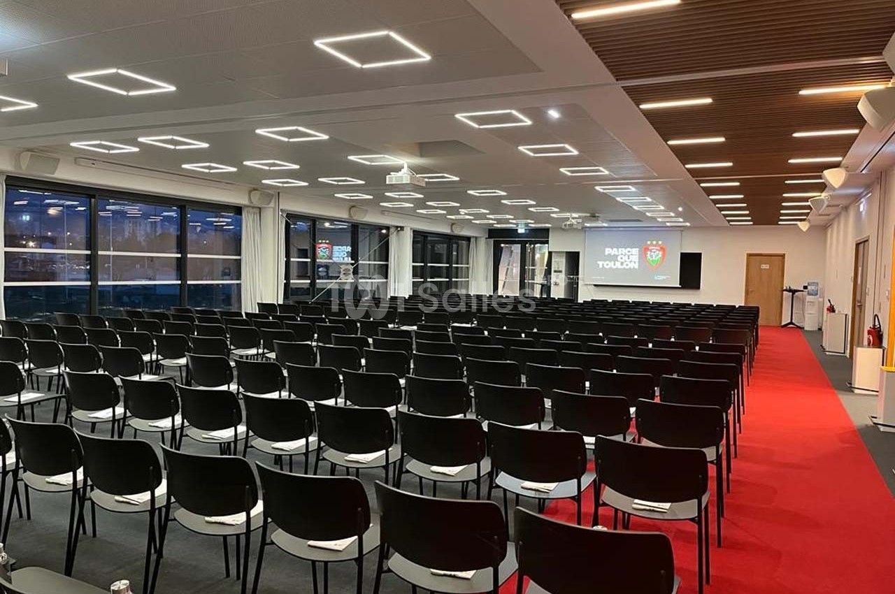 Salle de conférence moderne avec rangées de chaises noires, écran de projection et éclairage au plafond.