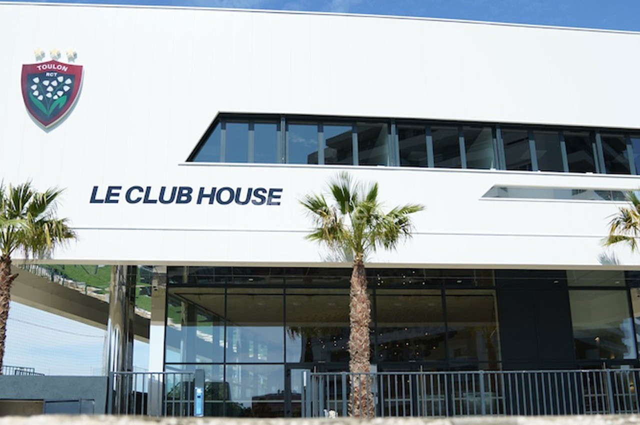 Façade moderne d'un bâtiment blanc avec l'inscription ’Le Club House’, des palmiers et un ciel bleu en arrière-plan.