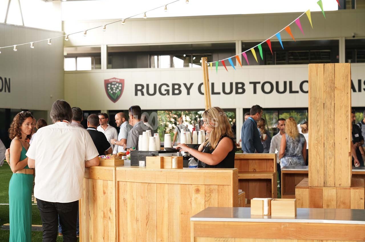 Des personnes discutent près de stands en bois dans un espace extérieur devant un bâtiment du Rugby Club Toulonnais.
