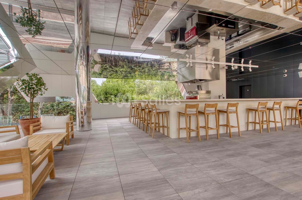 Salle moderne avec bar en bois, tabourets alignés, mobilier d'extérieur et grandes baies vitrées donnant sur la verdure.