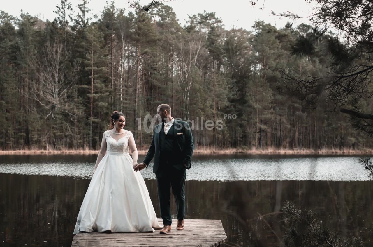 Un couple en tenue de mariage se tient sur un ponton en bois devant un lac entouré d'arbres.