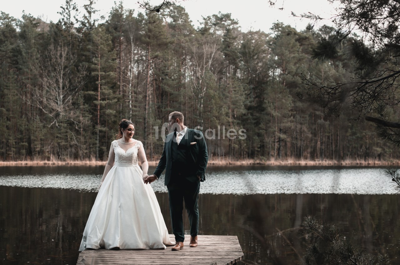 Un couple en tenue de mariage se tient sur un ponton en bois devant un lac entouré de forêt.