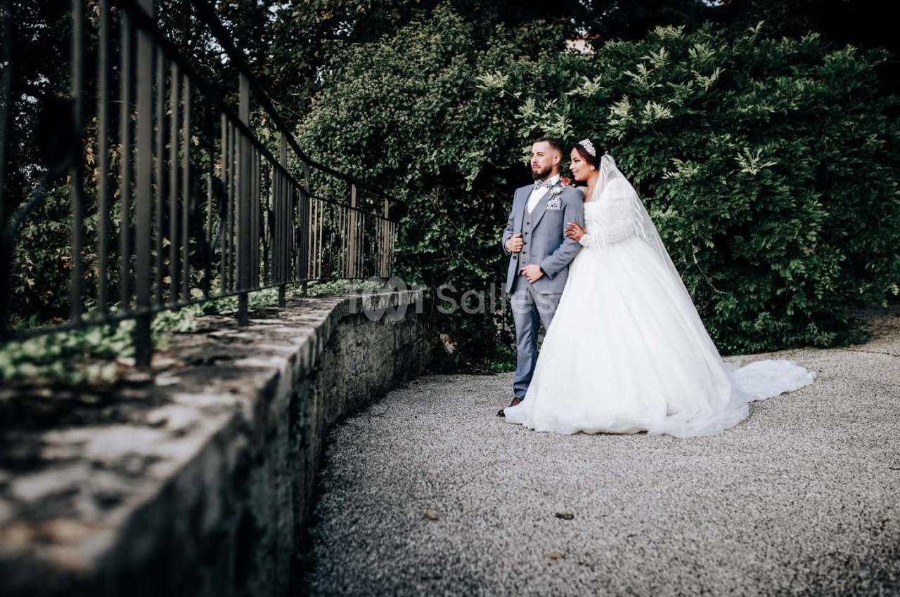 Un couple en tenue de mariage pose près d'une rambarde, entouré de verdure.