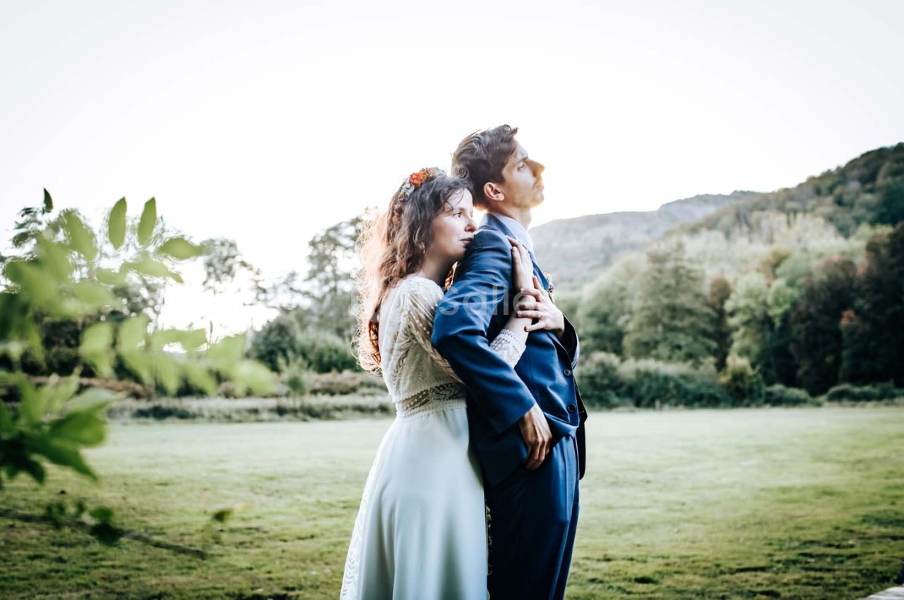 Un couple debout dans un champ verdoyant, la femme en robe blanche tenant l'homme en costume bleu.