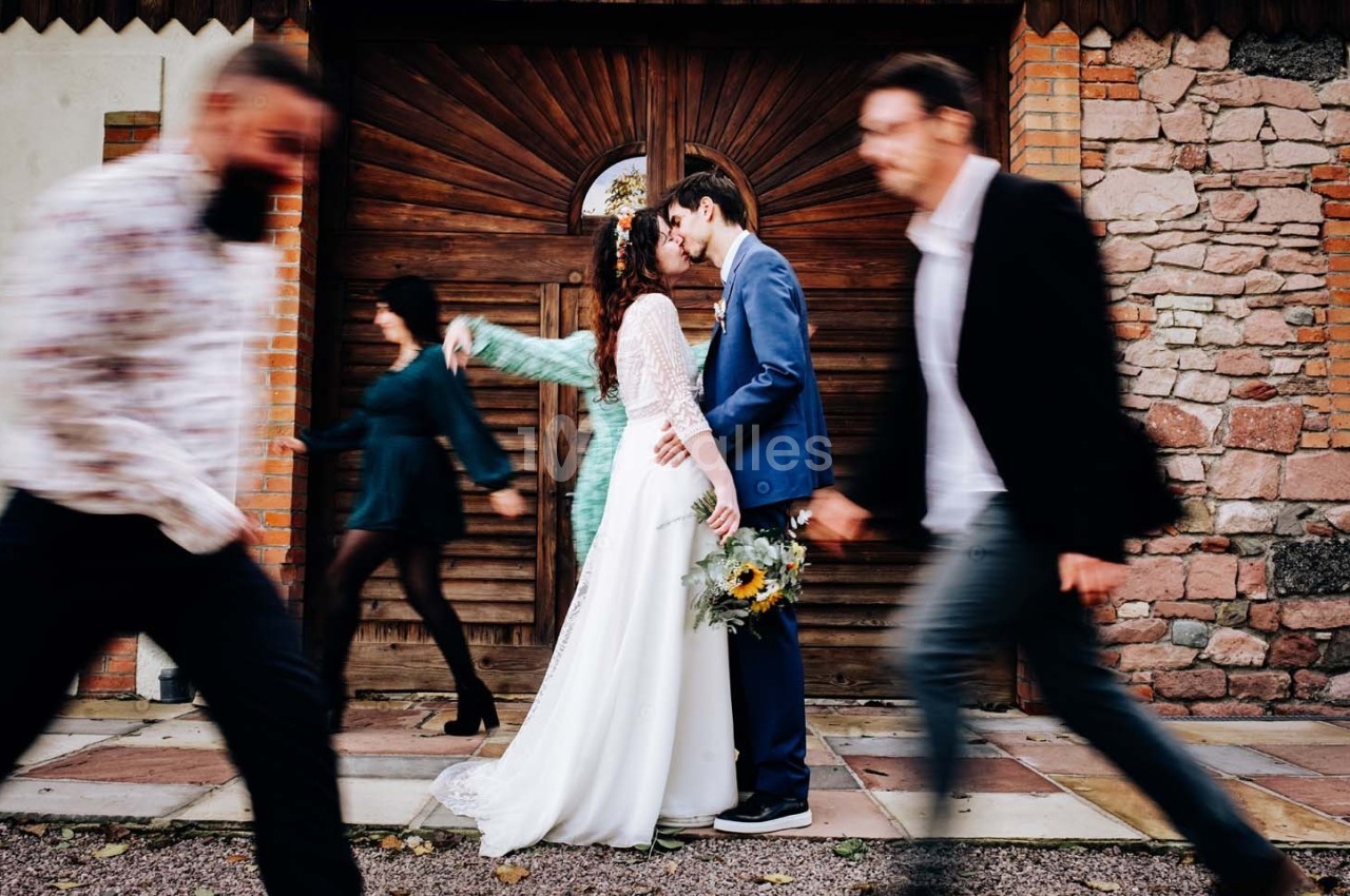 Un couple en tenue de mariage s'embrasse devant une porte en bois, des passants flous traversent la scène.