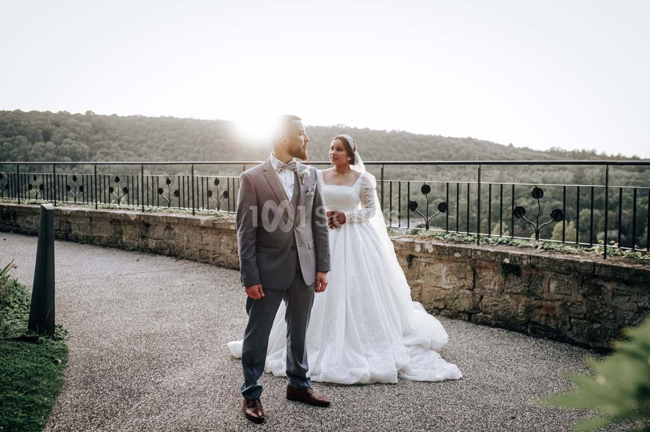 Un couple en tenue de mariage pose devant un paysage naturel avec une lumière du soleil en arrière-plan.