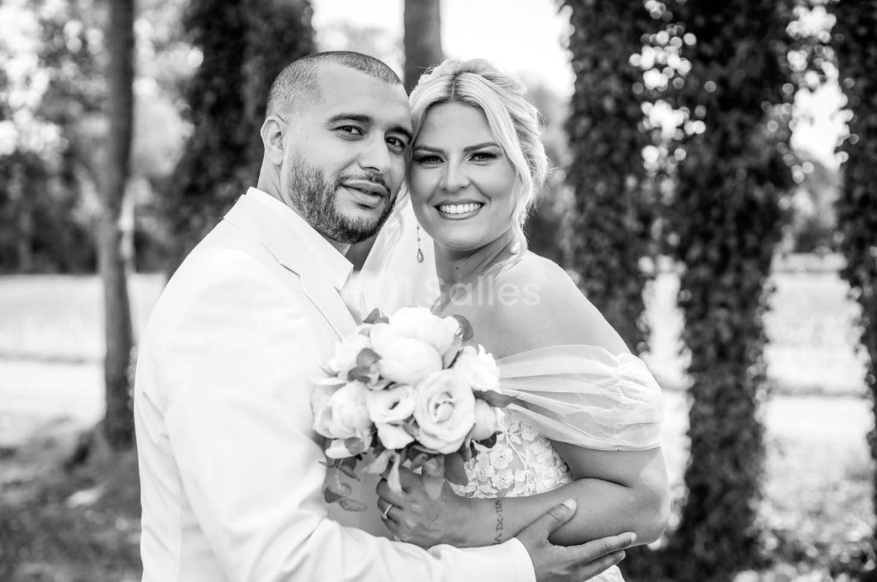 Un couple en tenue de mariage pose en extérieur, la mariée tenant un bouquet de fleurs.
