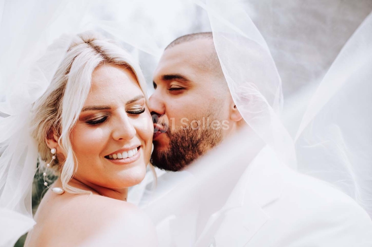 Un couple habillé en tenue de mariage, l'homme embrassant doucement la joue de la femme souriante sous un voile blanc.