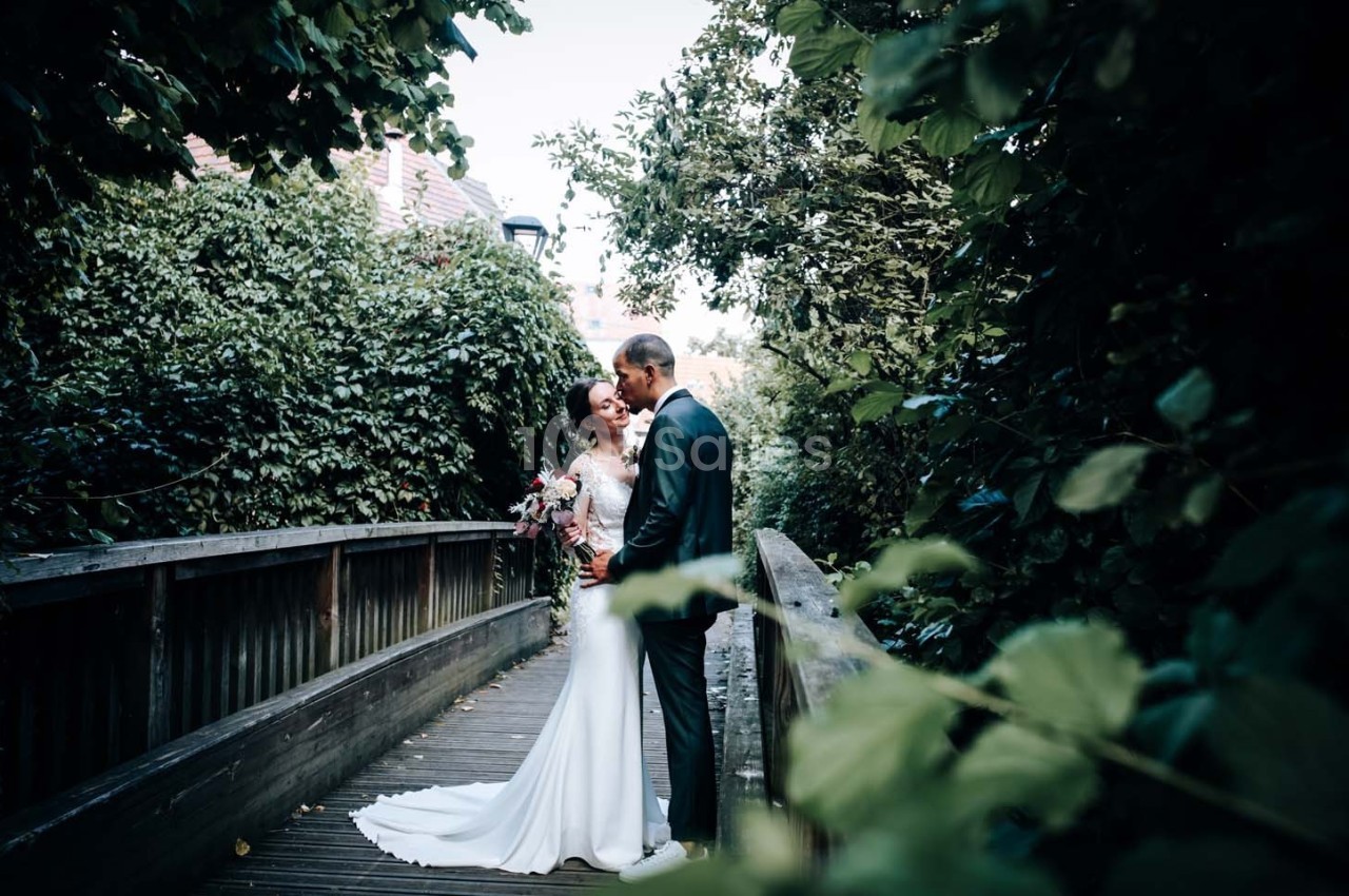 Un couple en tenue de mariage se tient sur un pont en bois entouré de végétation dense.