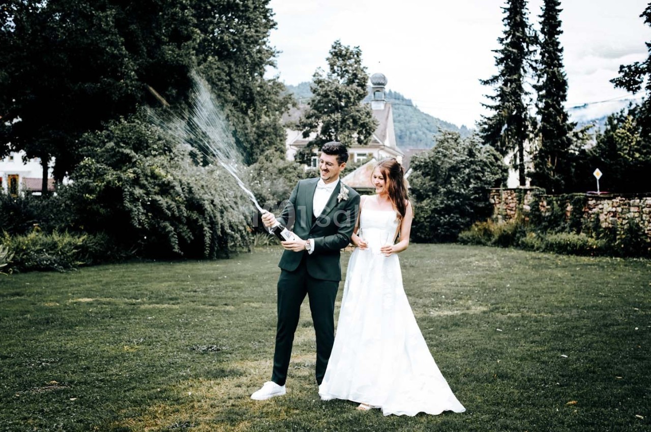 Un couple en tenue de mariage se tient sur une pelouse, l'homme arrosant avec un tuyau d'arrosage.