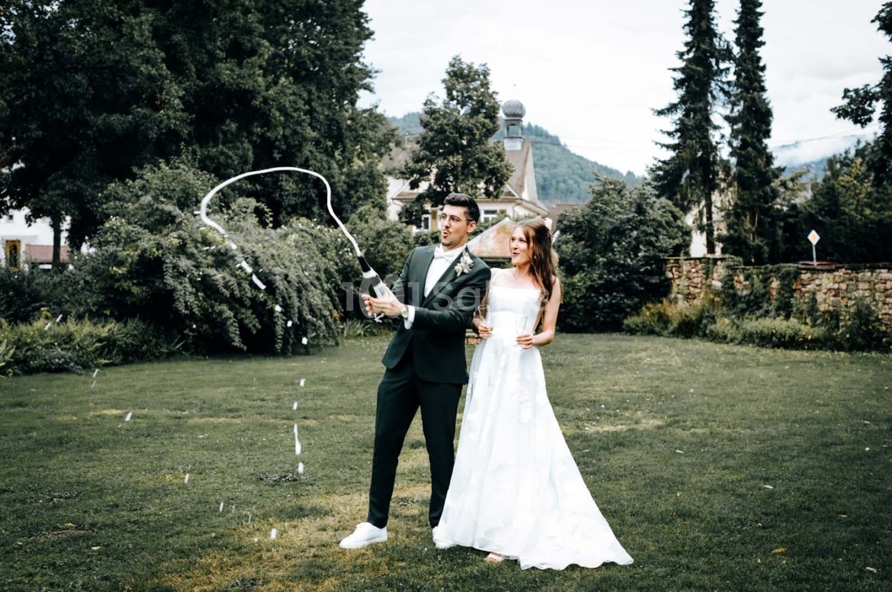 Un couple en tenue de mariage dans un jardin, l'homme ouvrant une bouteille de champagne qui éclabousse.