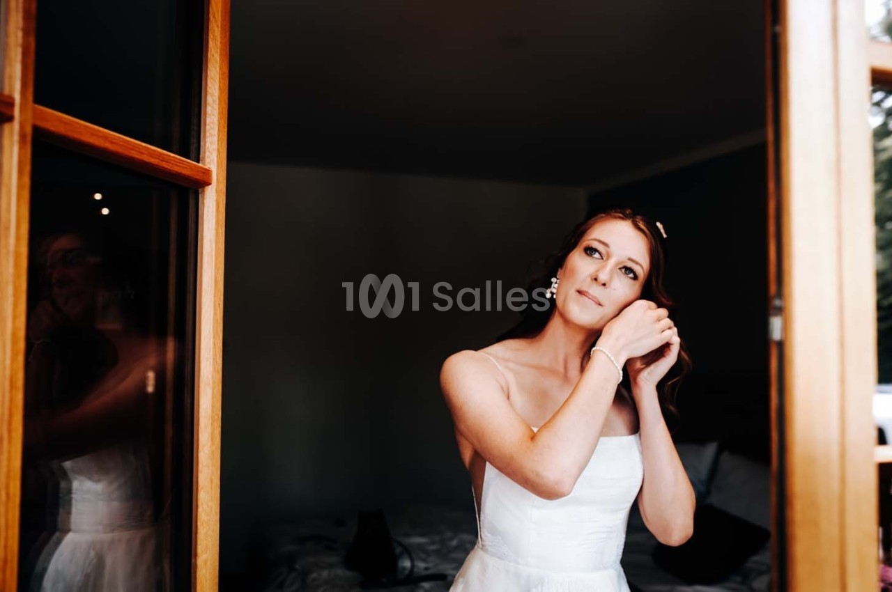 Une femme en robe blanche ajuste ses boucles d'oreilles devant une fenêtre ouverte dans une pièce sombre.