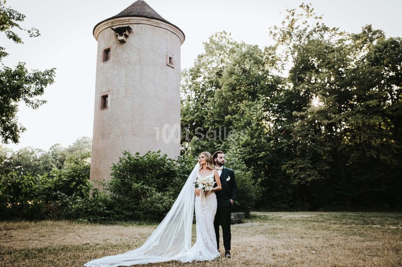 Un couple en tenue de mariage pose devant une tour en pierre entourée de végétation.