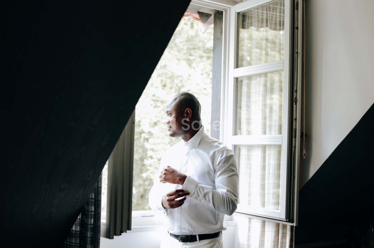 Homme en chemise blanche regardant par une fenêtre ouverte, baigné par une lumière naturelle.