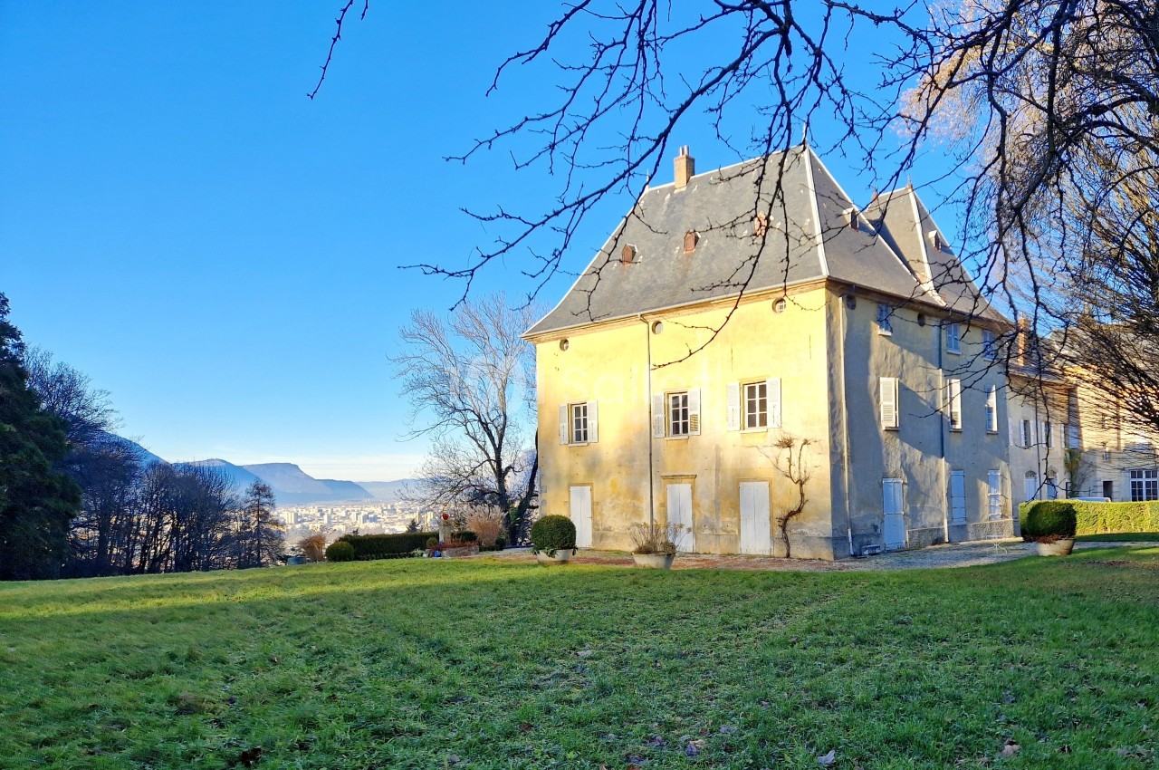 Grande maison ancienne avec toit en ardoise, entourée d'un jardin et d'arbres, sous un ciel bleu clair.