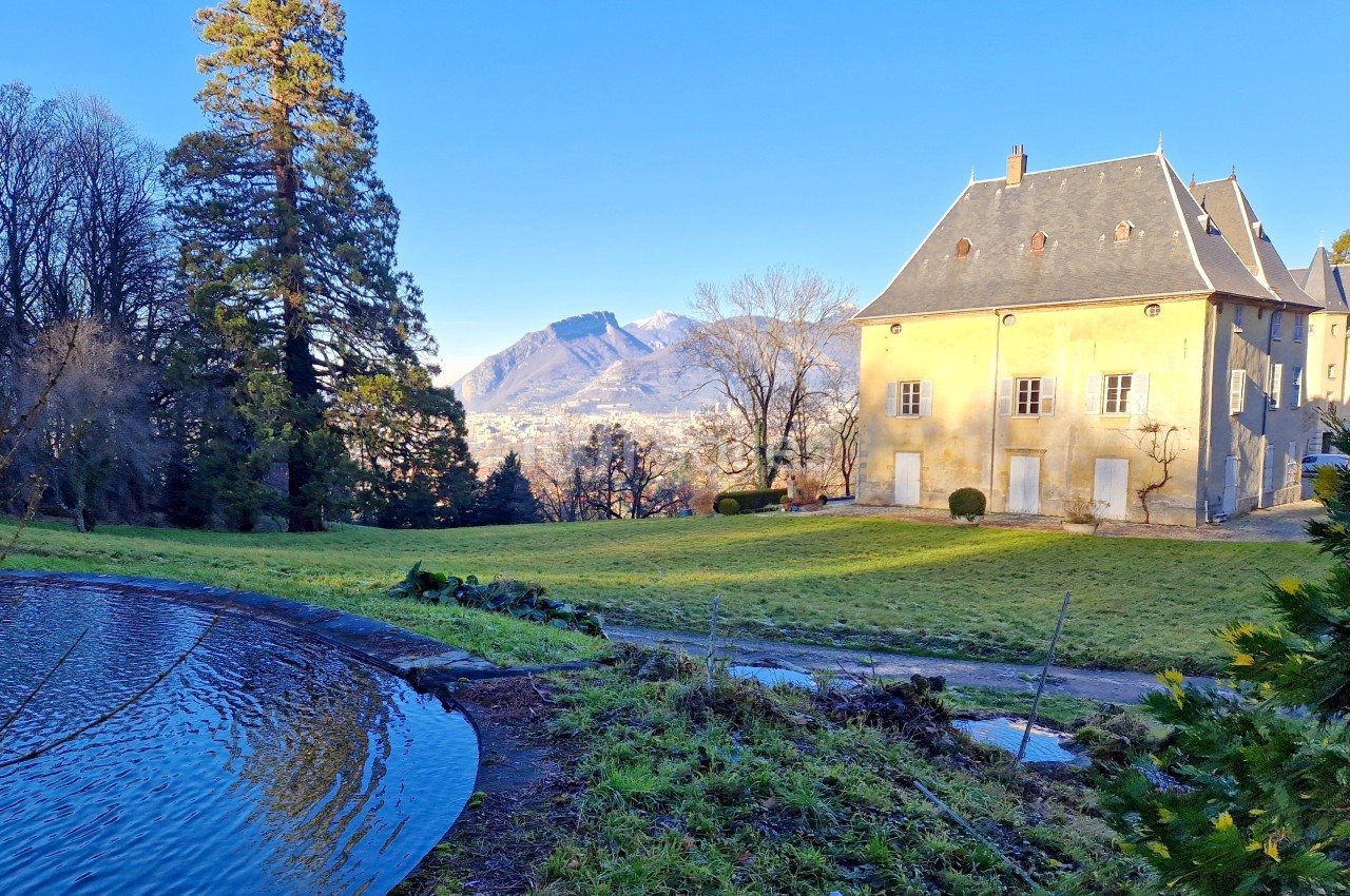 Vue d'un bâtiment ancien entouré de verdure, avec un étang au premier plan et des montagnes en arrière-plan.
