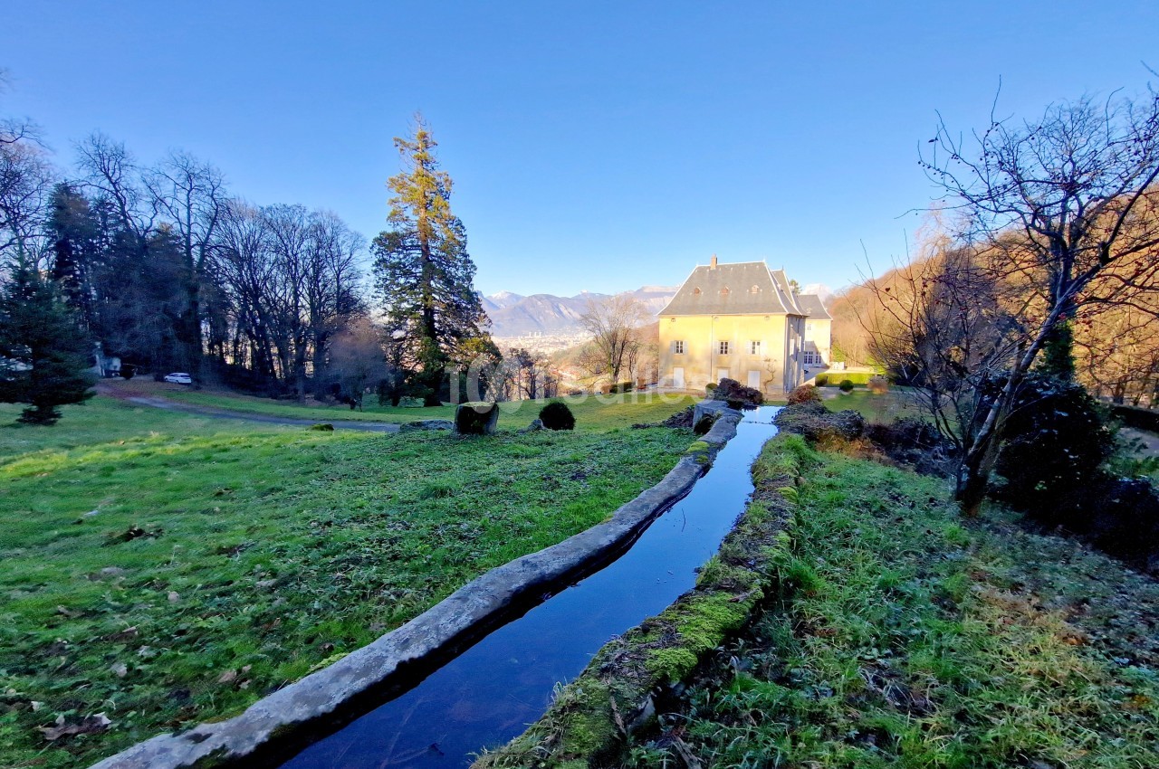 Manoir entouré d'un parc verdoyant avec un petit canal d'eau au premier plan, sous un ciel dégagé.
